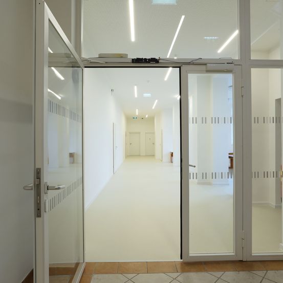 Ein leerer Korridor mit weißen Wänden, gefliestem Boden und zwei Glastüren. Eine Reihe von Lichtern verläuft entlang der Decke.