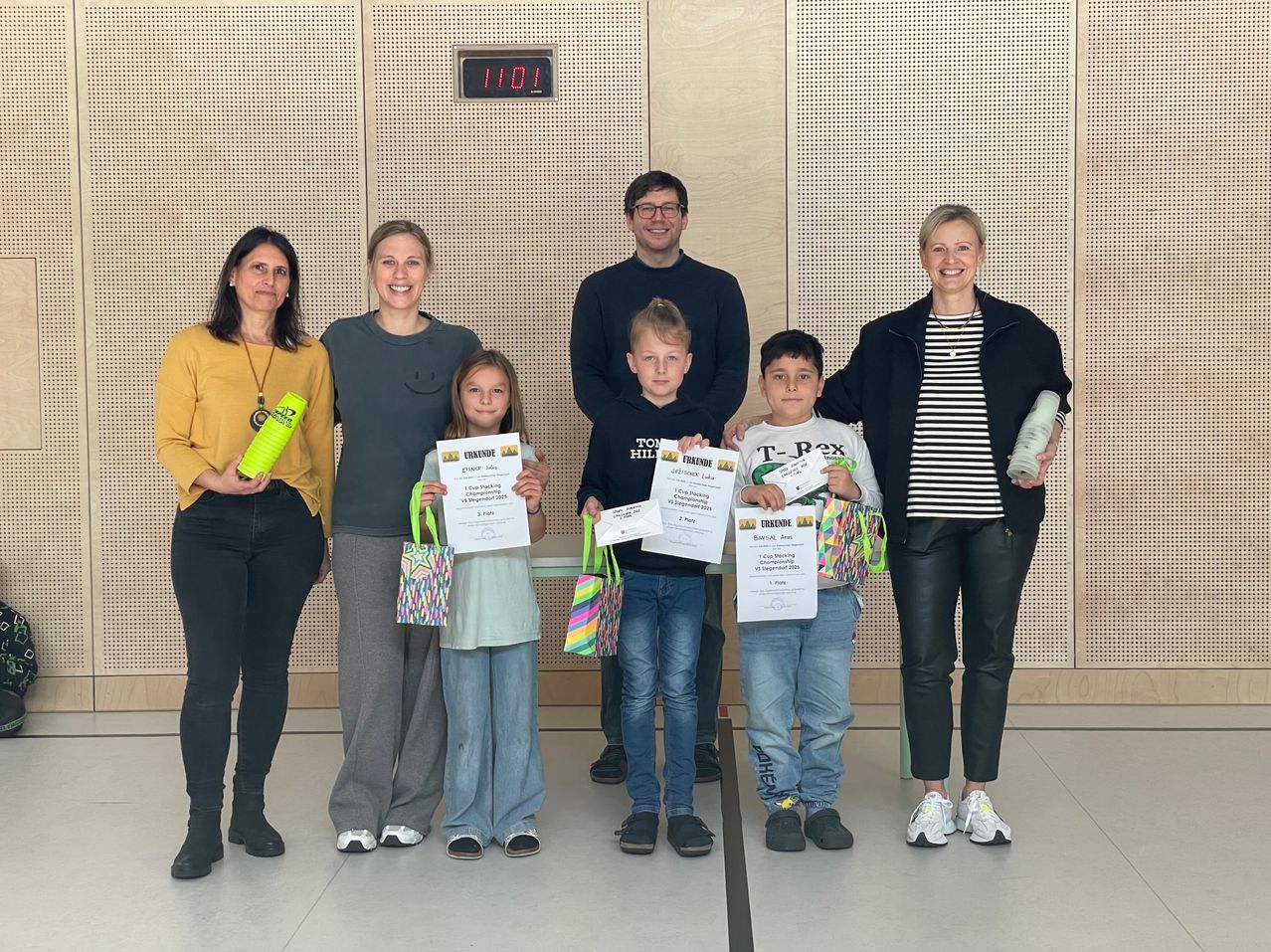 Eine Gruppe von Menschen, einschließlich Kindern und Erwachsenen, steht zusammen und hält Urkunden in der Hand und lächelt. Sie stehen vor einer weißen Wand mit einer digitalen Uhr, die 11:01 anzeigt.