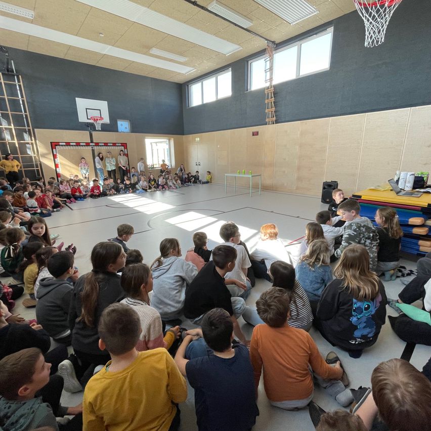 Hallenbasketballplatz, eine Gruppe von Schülern sitzt auf dem Boden und hört einer Präsentation zu, ein Tisch mit Laptop und Basketballkorb im Hintergrund.