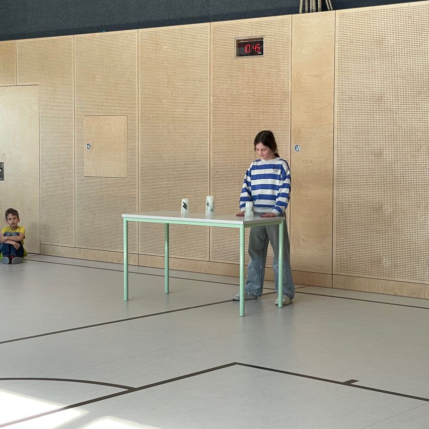 Ein junges Mädchen steht hinter einem Tisch in einem Raum mit Holzpaneelen und einer digitalen Uhr, die 10:45 anzeigt. Ein Junge sitzt vor dem Tisch auf dem Boden.