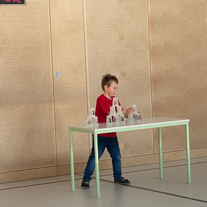 Ein Junge in einem roten Shirt und blauen Jeans versucht, Becher auf einem Tisch mit grünen Beinen zu stapeln.