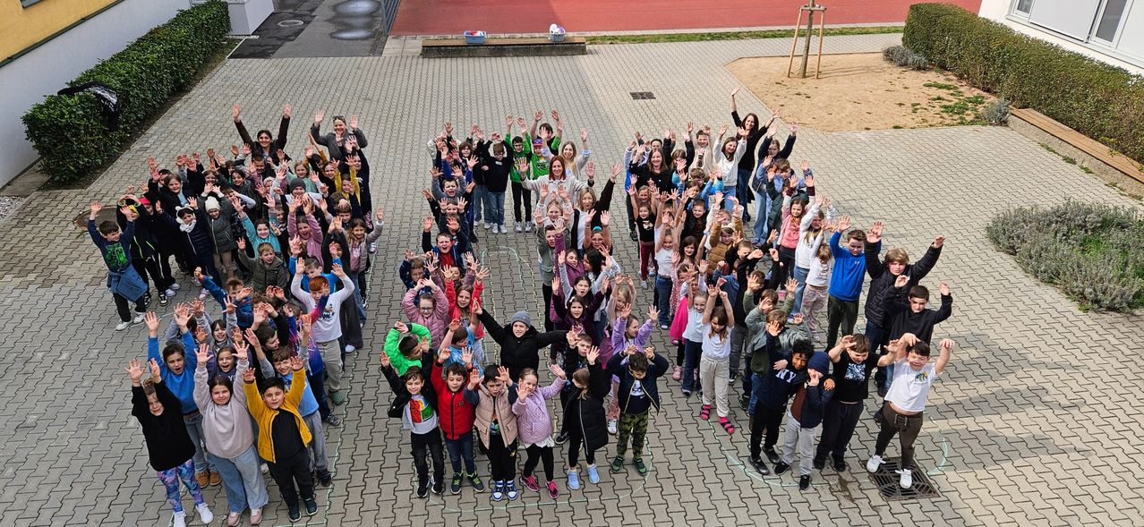 Eine große Gruppe von Kindern und Erwachsenen in Winterkleidung steht im Halbkreis auf einem gepflasterten Boden. Sie winken mit den Händen zur Kamera. Der Boden hat gezogene Linien. Ein rotes Feld ist im Hintergrund.