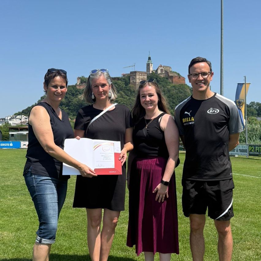Vier Personen posieren für ein Foto auf einem Rasenfeld mit einer Burg im Hintergrund. Eine hält ein Zertifikat, und alle lächeln.