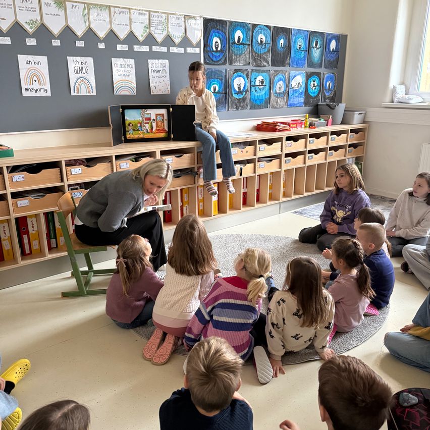 Eine Lehrerin liest jungen Kindern in einem Klassenzimmer eine Geschichte vor. Die Kinder sitzen im Kreis auf einem Teppich. Ein Kind sitzt auf einem Stuhl und ein anderes auf einem Regal. Ein Monitor und einige Bücher sind auf dem Regal.