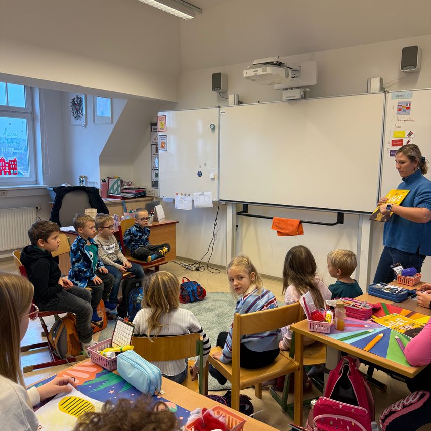 Ein Klassenzimmer mit Kindern, die auf Stühlen sitzen, und einer Lehrerin, die ein Buch liest. Eine Tafel und ein Projektor sind an der Wand montiert. Kinder beschäftigen sich mit Aktivitäten an ihren Schreibtischen.