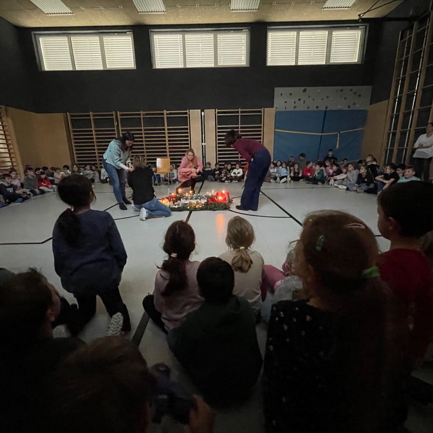 Eine Gruppe von Kindern und zwei Erwachsenen versammelt sich in einer Turnhalle für eine Veranstaltung. Zwei Erwachsene richten Kerzen auf dem Boden an.