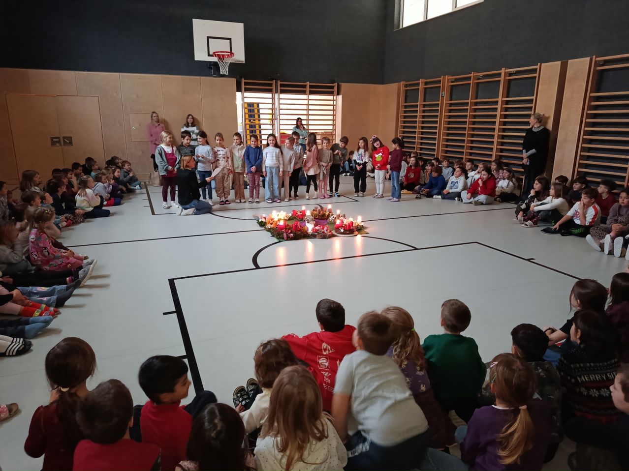 In einer Turnhalle steht eine Gruppe von Kindern um eine kerzenbeleuchtete Anordnung. Eine Person liest von einem Papier, und andere sitzen auf dem Boden. Ein Basketballkorb ist an der Wand zu sehen.