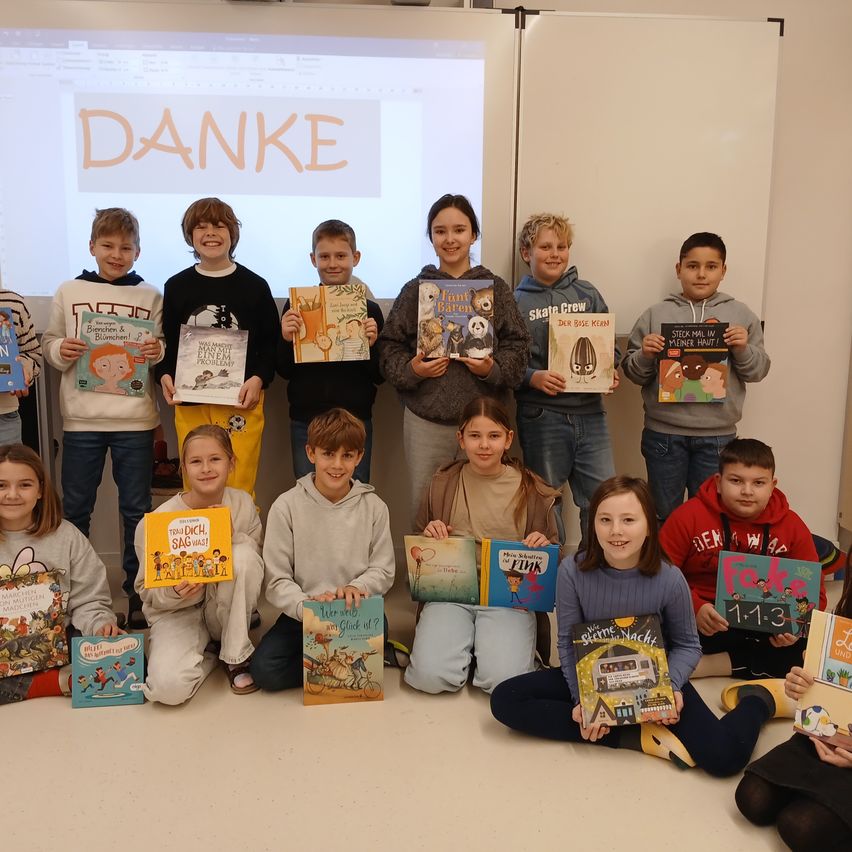 Eine Gruppe von Kindern in einem Klassenzimmer posiert für ein Foto, während sie Bücher halten. Sie sitzen in zwei Reihen, mit einer Projektionsleinwand hinter ihnen, auf der das Wort Danke steht.