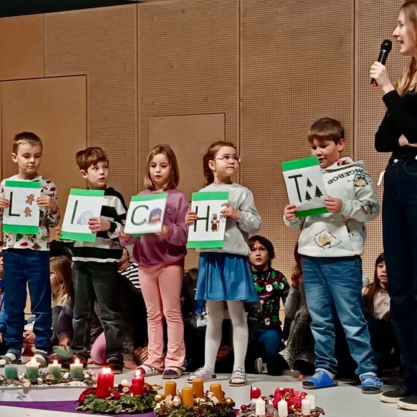 Kinder auf der Bühne halten Buchstaben, die ein Wort bilden. Eine Frau mit Mikrofon steht in der Nähe. Dahinter sitzen andere Kinder auf dem Boden. Die Bühne ist mit Kerzen und Grünzeug geschmückt.