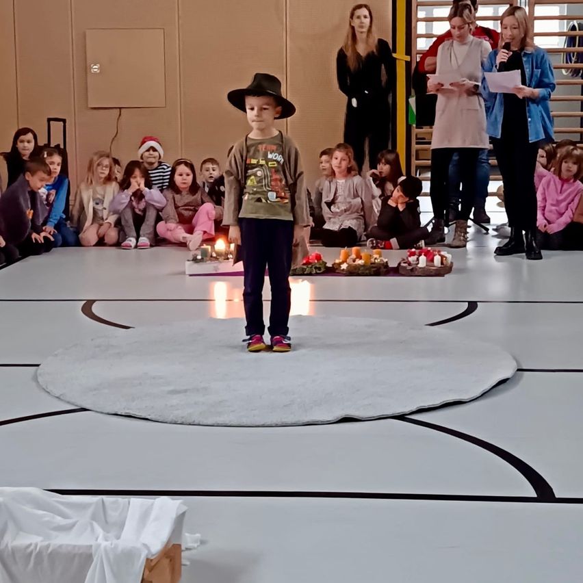 Eine Gruppe von Kindern sitzt auf dem Boden und beobachtet einen Jungen, der in der Mitte eines Kreises steht. Kerzen sind vor ihm auf dem Boden angeordnet. Zwei Frauen stehen hinter dem Jungen und halten Papiere.