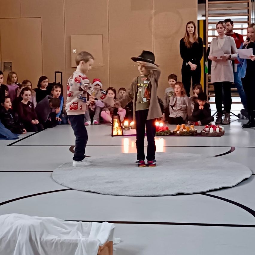 Zwei Kinder stehen auf einem runden Teppich in einem Raum mit Kerzen, umgeben von einem sitzenden Publikum. Erwachsene stehen hinten, einer hält ein Mikrofon.