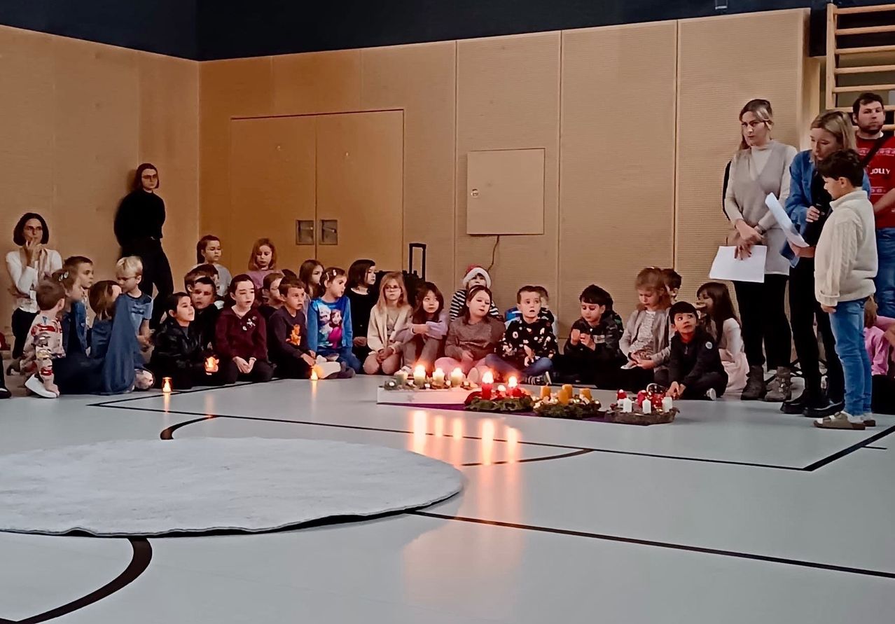 Kinder und Erwachsene sitzen in einem Kreis um eine zentrale Anordnung von Kerzen, wahrscheinlich an einer zeremoniellen Veranstaltung teilnehmend. Der Raum hat beige Wände und einen gefliesten Boden.