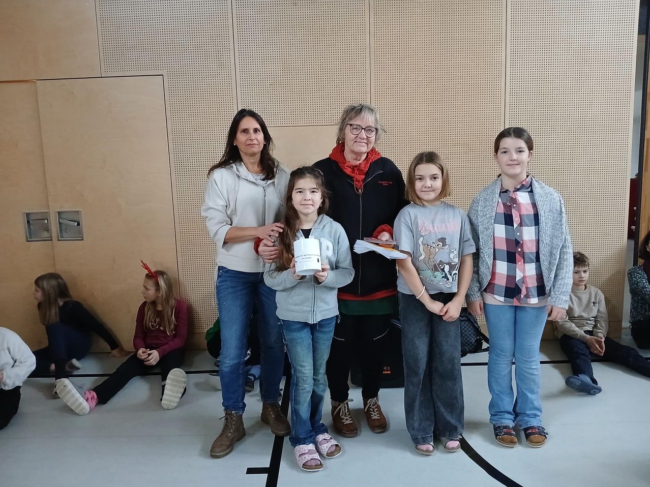 Eine Gruppe von Frauen und Mädchen steht zusammen für ein Foto. Das Mädchen in der Mitte hält einen Becher. Sie sind in einem Raum mit einer beigen Wand.