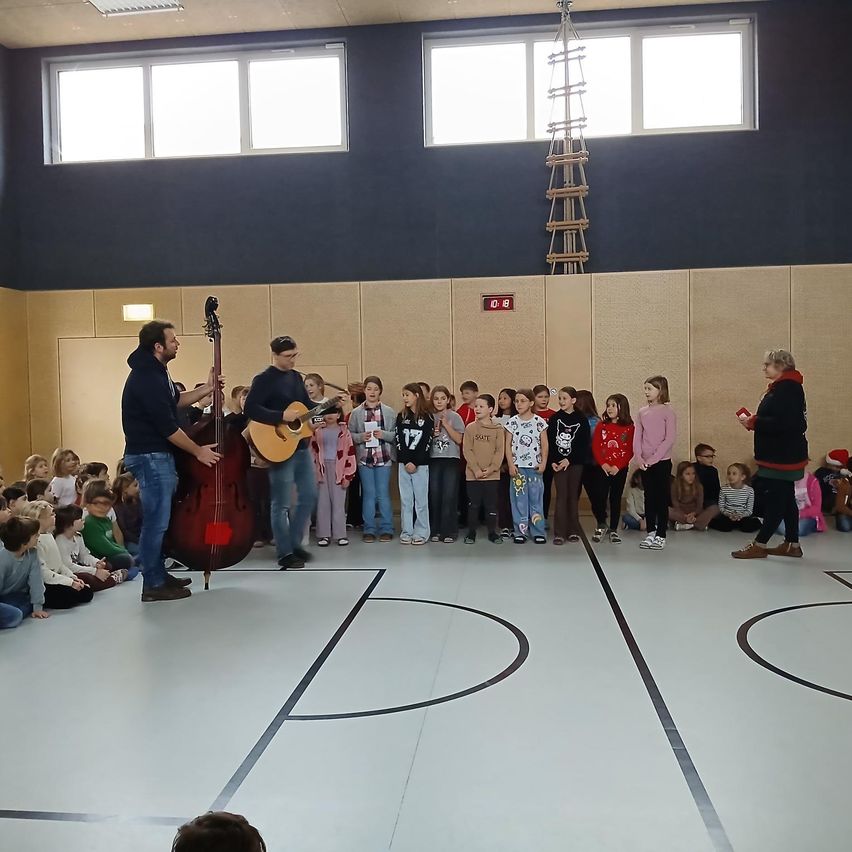 Eine Gruppe von Kindern steht in einer Reihe und beobachtet zwei Männer, die in einer Turnhalle Musikinstrumente spielen.
