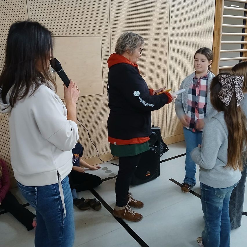Eine Frau mit einem Mikrofon steht vor einer Gruppe von Menschen. Eine ältere Frau in einer Kapuzenjacke reicht einem Mädchen ein Papier. Der Ort ist drinnen mit einer Wand im Hintergrund.