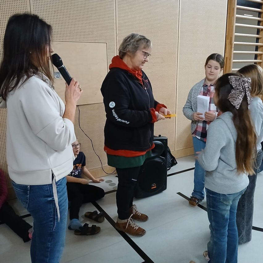 Eine Gruppe von Menschen ist in einem Raum. Eine Frau hält ein Mikrofon und eine andere Frau hält eine gelbe Karte. Eine dritte Frau liest ein Papier.