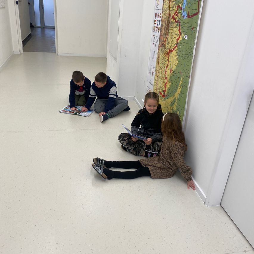 Vier Kinder sitzen auf dem Boden und lesen ein Buch. Sie sind in einem Raum mit weißen Wänden und einer Karte an der Wand. Der Boden ist glänzend und sauber.