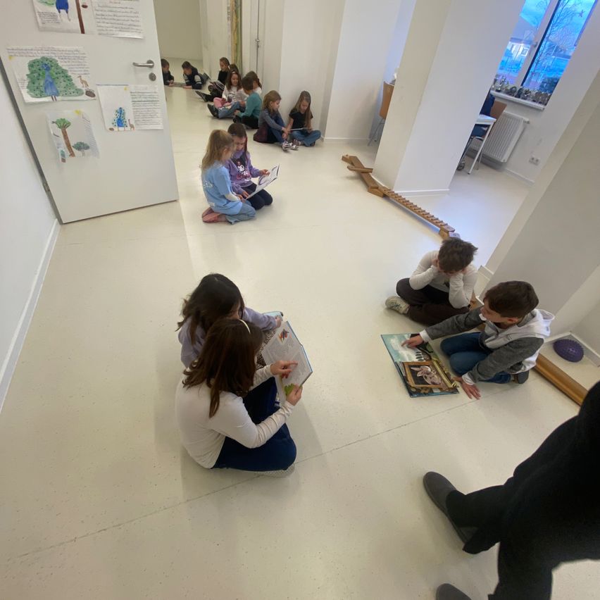 Mehrere Kinder sitzen auf dem Boden und lesen Bücher, einige haben Bücher auf dem Schoß offen. Eine Holzleiter liegt auf dem Boden. Eine Person steht in der Nähe.