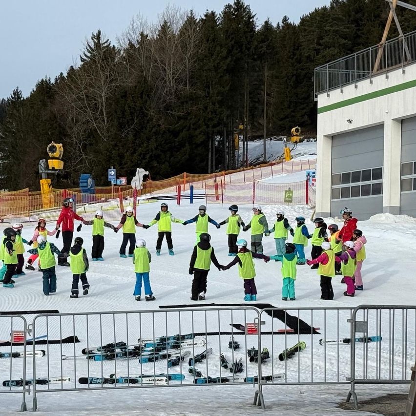 Eine Gruppe von Kindern in gelben Westen halten sich an den Händen in einem Kreis auf einer verschneiten Oberfläche. Vor ihnen liegen Skier und andere Ausrüstung. Dahinter befinden sich Zäune und ein Gebäude mit Balkon. Bäume und Schnee sind im Hintergrund.