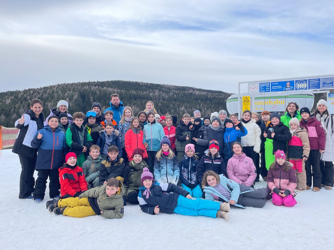 Eine Gruppe von Kindern und Erwachsenen in Winterkleidung posiert für ein Foto auf einem verschneiten Berg mit einem Schild dahinter.