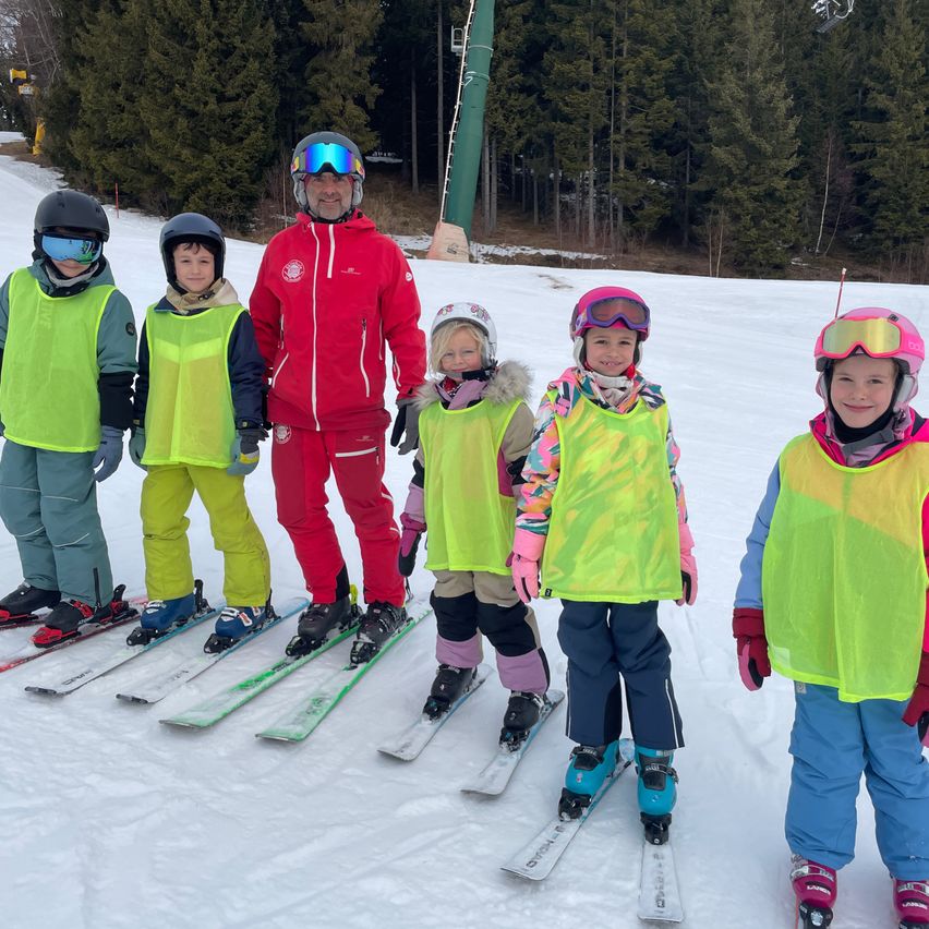 Fünf Kinder in Skiausrüstung stehen auf einer verschneiten Piste, mit einem Mann in einem roten Skianzug in der Mitte. Bäume und eine Skiliftanlage sind im Hintergrund.