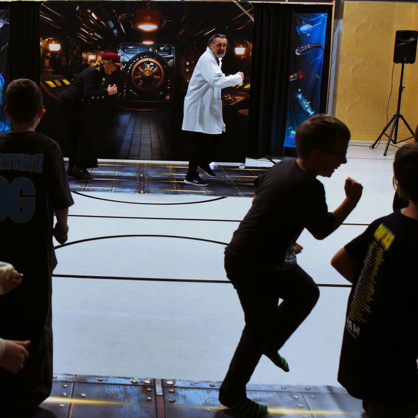 Eine Gruppe von Kindern in schwarzen T-Shirts spielt ein Spiel auf einer Bühne mit einem Mann im Laborkittel und einer Frau in Uniform. Die Bühne hat eine große Leinwand und ein großes Rad im Hintergrund.