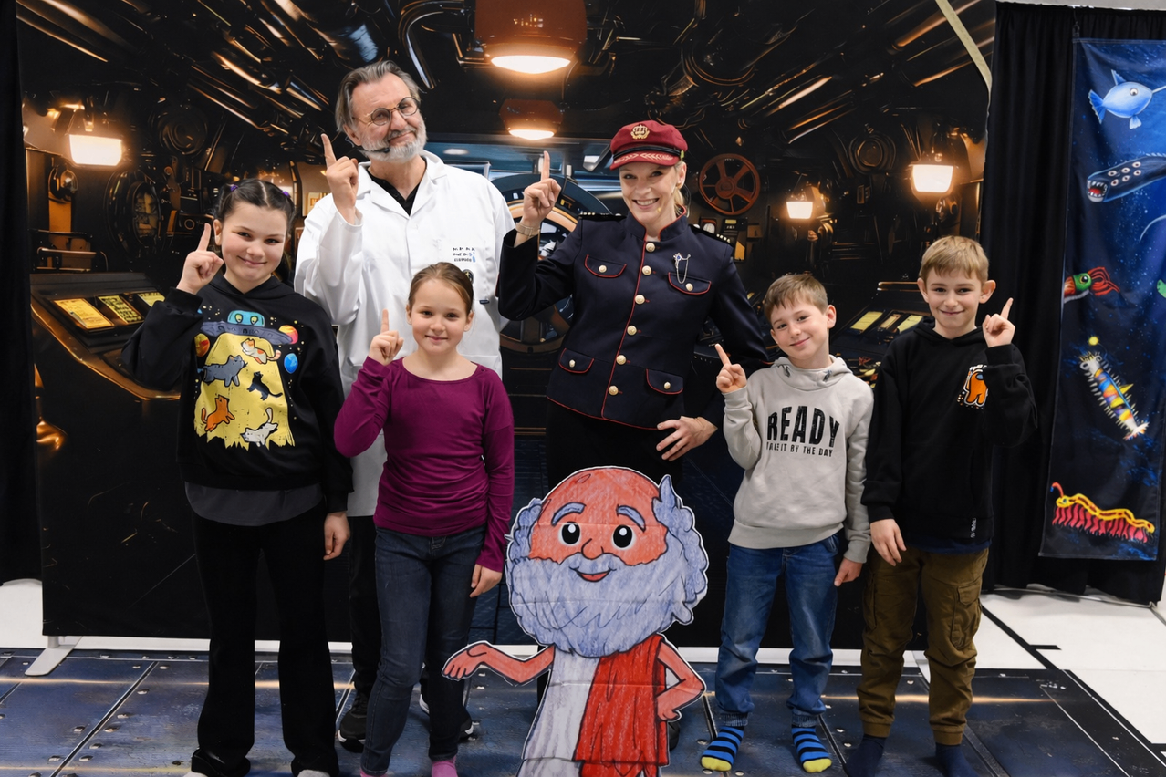 Eine Gruppe von Kindern und zwei Erwachsenen stehen zusammen in einem Raum. Sie scheinen für ein Foto zu posieren. Die Kinder zeigen mit den Fingern. Der Erwachsene auf der linken Seite ist in einem weißen Mantel gekleidet.