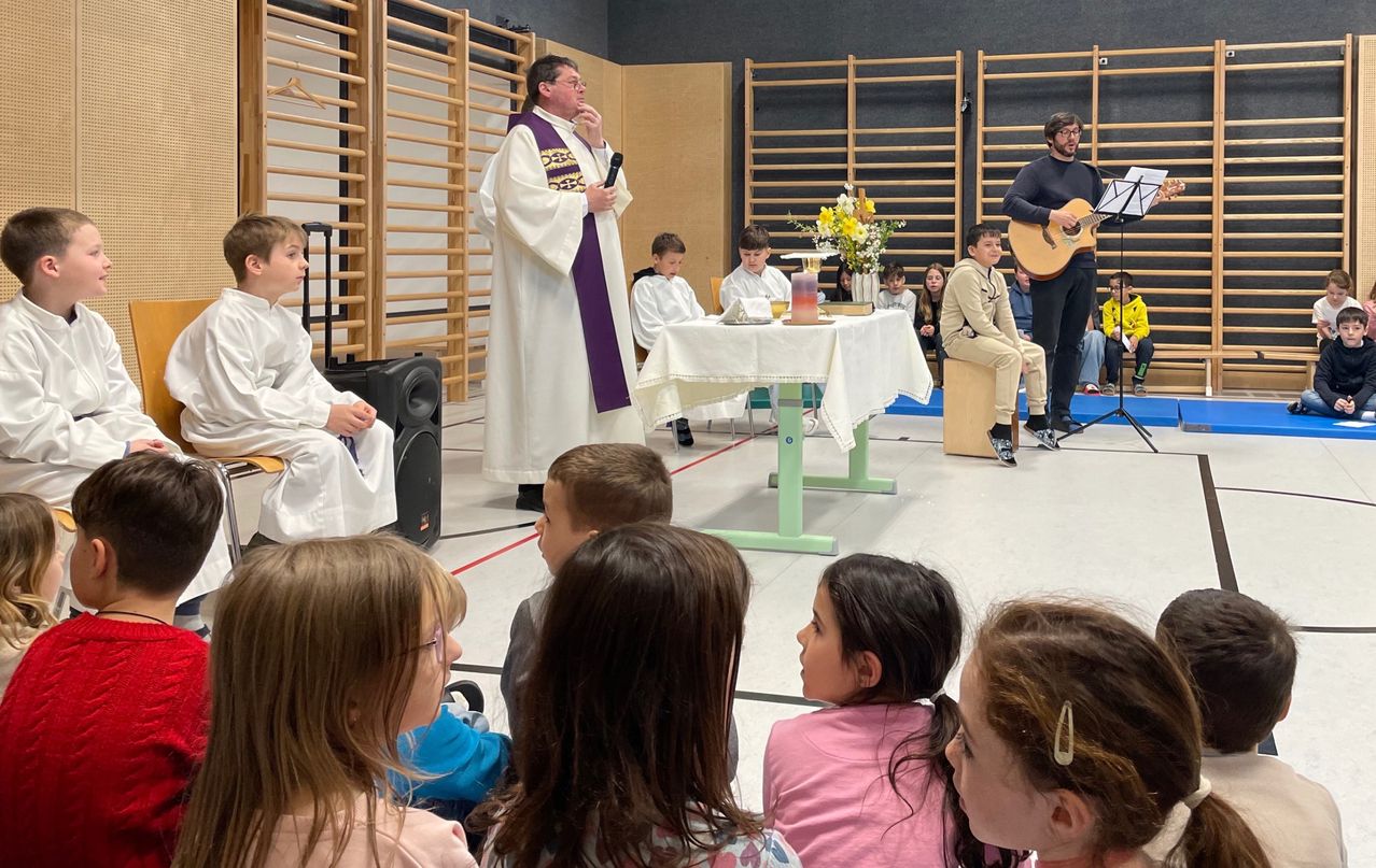 Ein Priester spricht eine Gruppe von Kindern an, während ein Mann Gitarre spielt. Der Priester hält ein Mikrofon, und die Kinder sitzen aufmerksam zu.