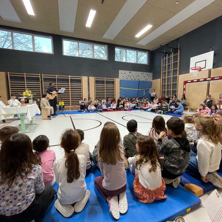 Ein Mann spielt Gitarre auf einer Bühne, während eine Menge Kinder und Erwachsene zuhören. Die Bühne befindet sich in einer Turnhalle mit einem Basketballplatz und großen Fenstern.