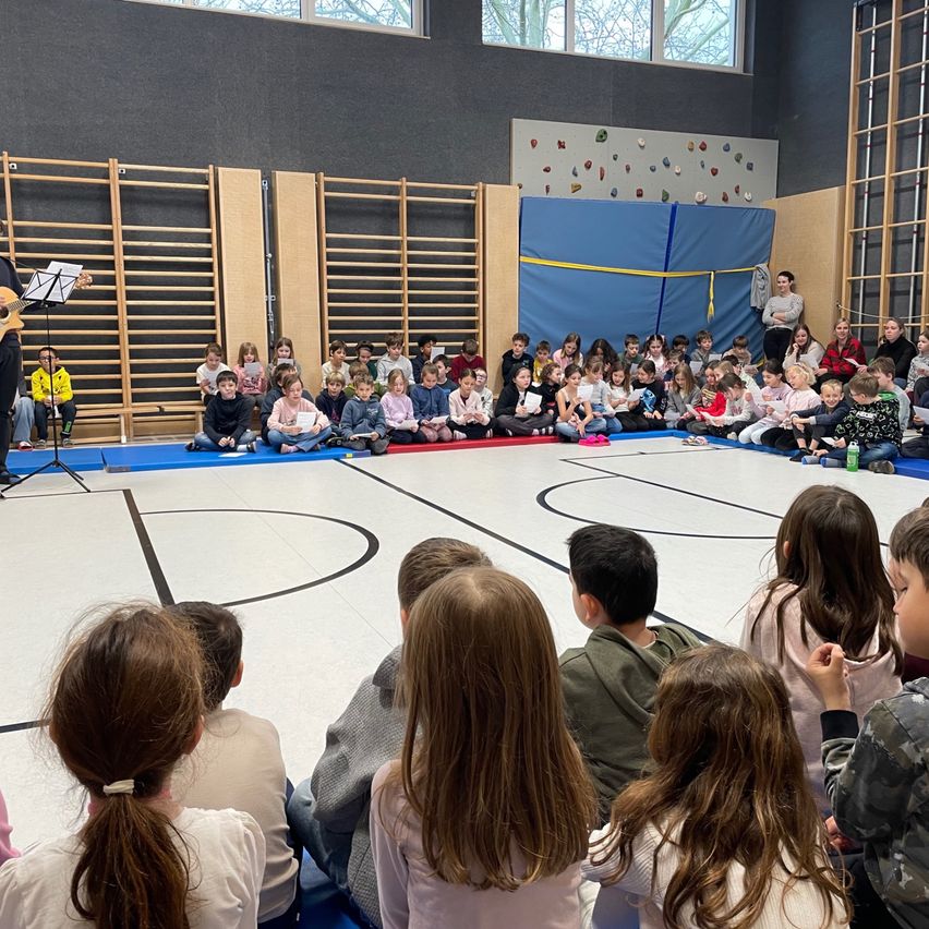 Kinder sitzen in einem Kreis um einen Mann, der auf einer Bühne in einer Turnhalle eine Gitarre spielt. Sie schauen ihm aufmerksam zu.