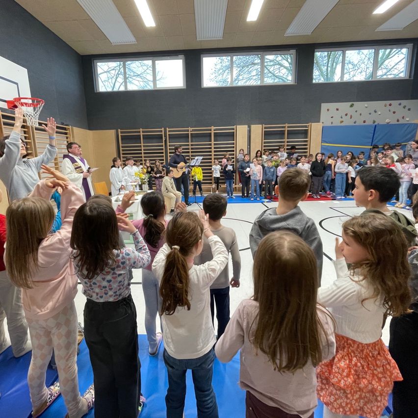 Kinder versammeln sich in einer Turnhalle, einige stehen auf einer blauen Matte. Ein Mann spielt Gitarre, während andere singen. Ein Priester hält einen Basketballkorb.