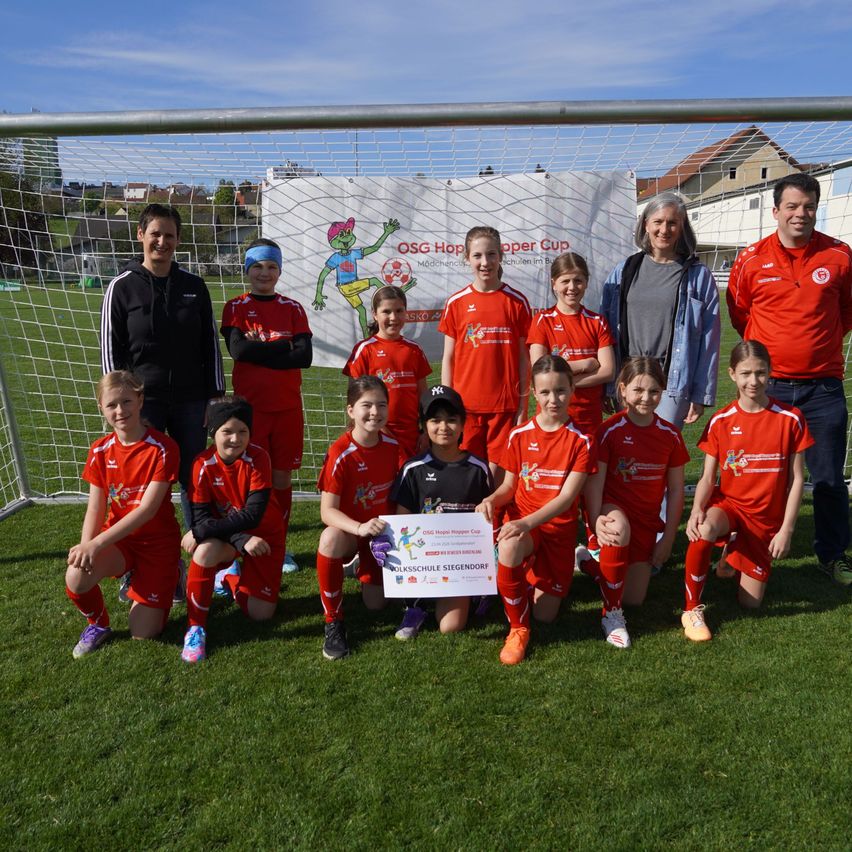 Eine Jugendfußballmannschaft posiert mit ihrem Pokal auf einem Fußballfeld. Sie tragen rote Trikots. Dahinter stehen zwei Erwachsene, und eine weiße Fahne ist zu sehen.