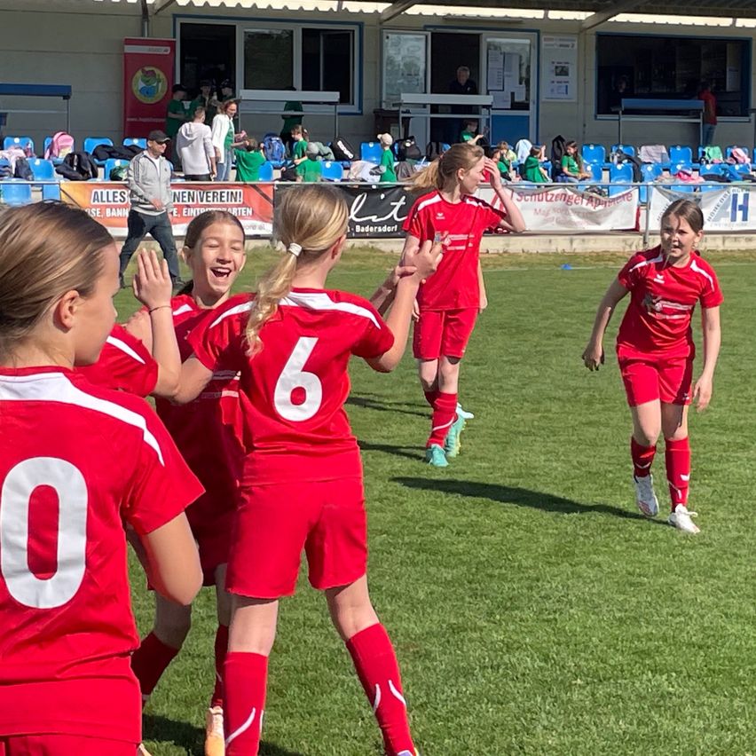 Eine Gruppe junger Mädchen in roten Fußballtrikots feiert auf dem Spielfeld, während der Hintergrund der Zuschauer verschwommen ist.
