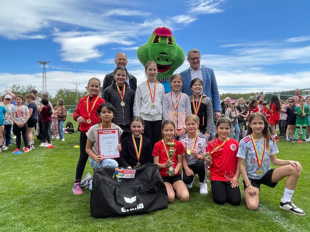 Eine Gruppe junger Mädchen und zwei Männer posieren für ein Foto. Sie halten Medaillen und Urkunden, und ein Mädchen hält einen Pokal. Sie stehen auf einem Rasenfeld.