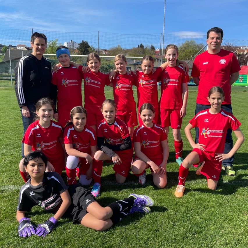 Eine Fußballmannschaft junger Mädchen posiert mit zwei Erwachsenen auf einem Rasenfeld mit einem Tor im Hintergrund.