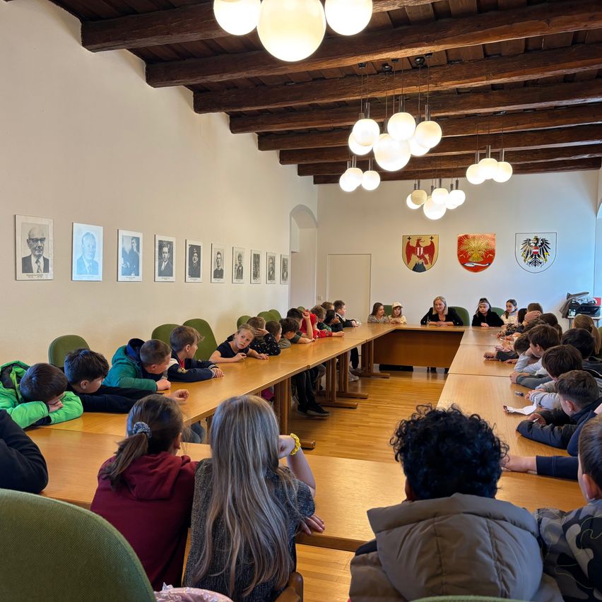 Eine Gruppe von Kindern sitzt an einem langen Tisch in einem Besprechungsraum, mit Erwachsenen am Kopfende. Gerahmte Bilder schmücken die Wand.