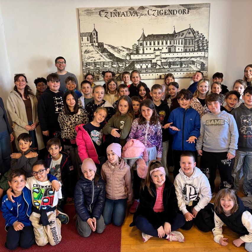 Eine Gruppe von Kindern und Erwachsenen posiert vor einem Banner mit einer Schlosszeichnung. Einige tragen Brillen, Mützen und Turnschuhe. Der Boden ist aus Holz.