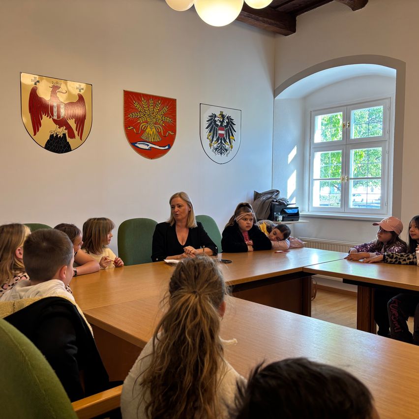 Eine Gruppe von Kindern sitzt um einen Konferenztisch herum mit einer Frau an der Spitze. Hinter ihr sind drei Schilder an der Wand angebracht.
