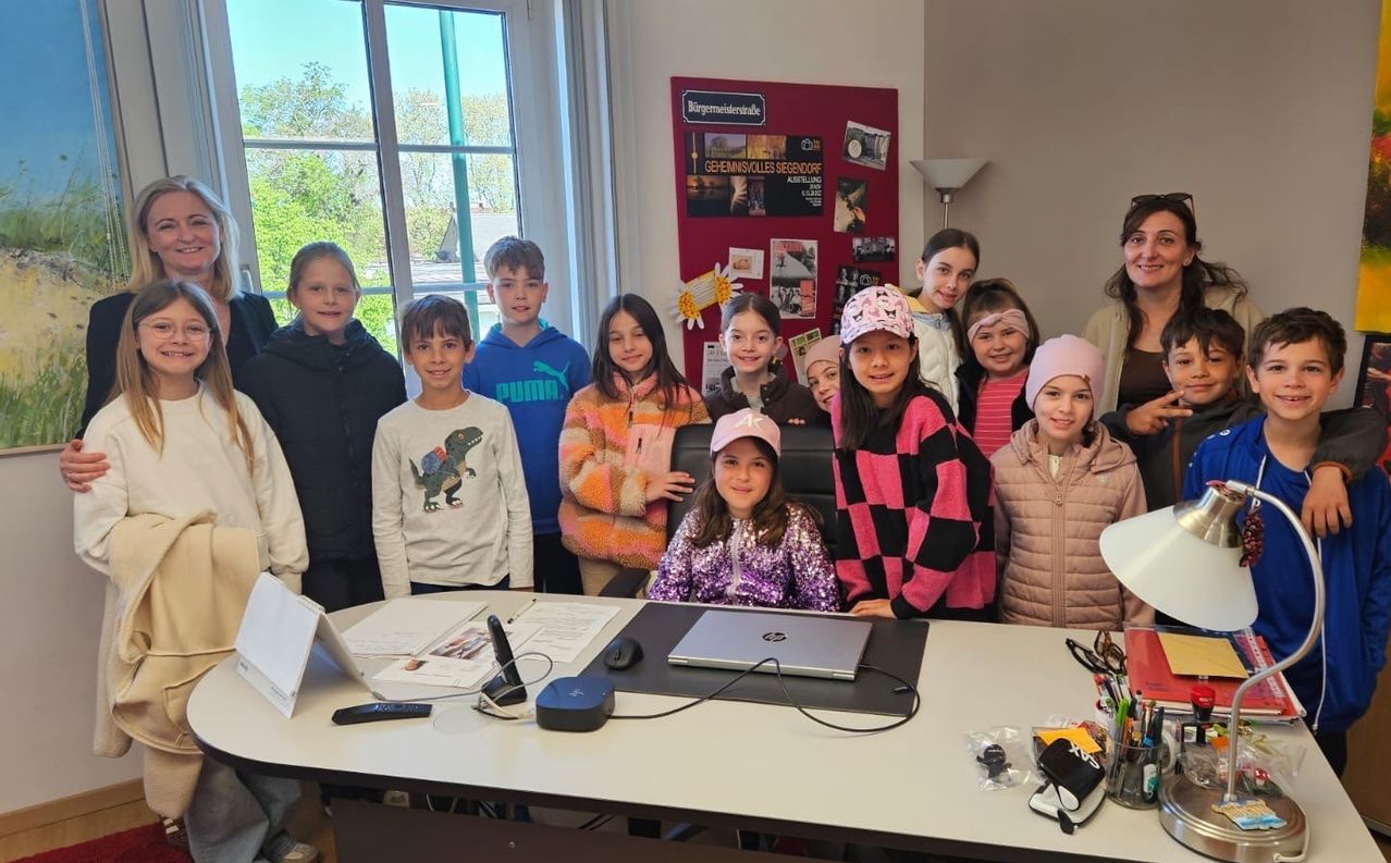 Eine Gruppe junger Schüler steht vor einem Schreibtisch mit Laptop und posiert für ein Foto. Im Hintergrund befindet sich eine Pinnwand mit verschiedenen Postern und einer Lampe.