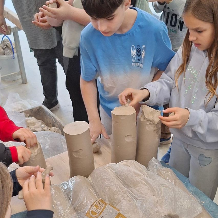 Mehrere Kinder sind um einen Tisch versammelt und arbeiten jeweils an Lehmskulpturen. Der Tisch ist mit Plastik bedeckt und darauf befinden sich Lehmstücke und Werkzeuge. Die Kinder tragen Freizeitkleidung.