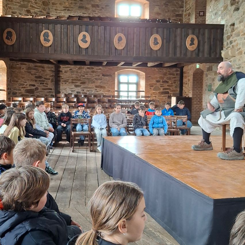 Ein Mann sitzt auf einer Bühne vor Kindern. Sie sitzen auf Holzstühlen. Die Bühne hat einen Holzboden und einen schwarzen Vorhang. Der Raum hat eine Steinwand und eine Holzbalkon.