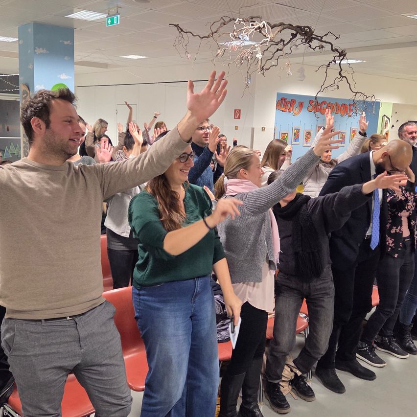 A group of people in an indoor area, raising their hands. The person in front is smiling. The room has a blue wall with decorations and a sign. The ceiling has lights and a branch with hanging objects.