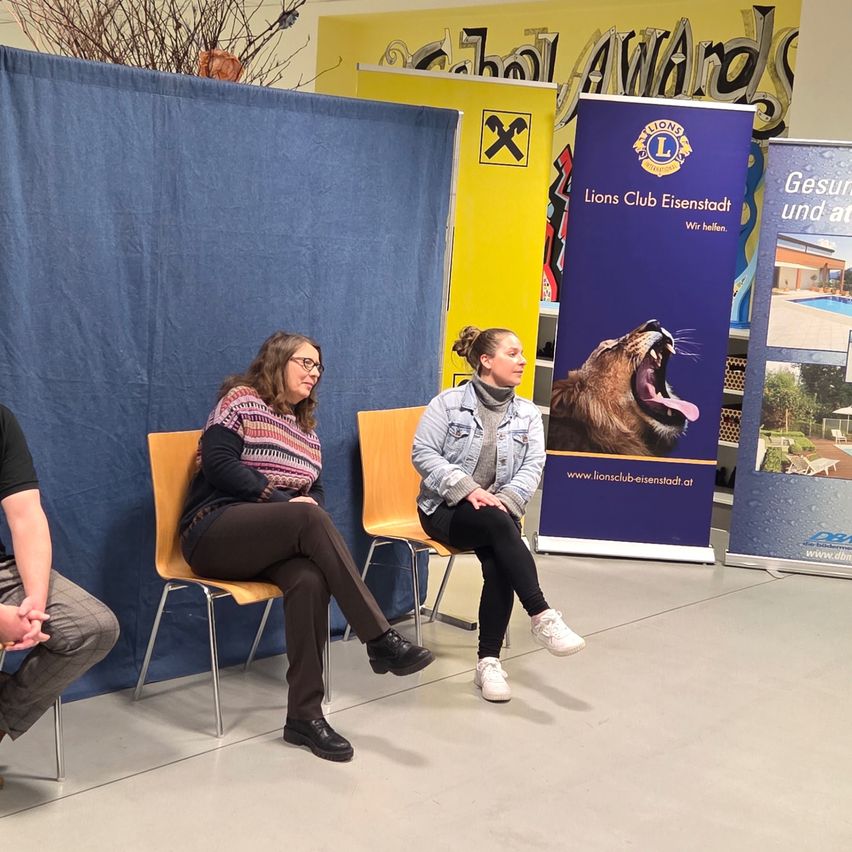 Three people sit in front of a backdrop. Two women in glasses and jeans sit in yellow chairs. A man in black jeans sits on a blue chair. A blue banner is behind them.