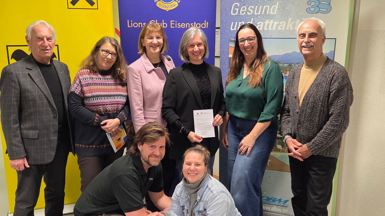A group of people are posing for a picture. A woman in a black blazer holds a certificate. Behind her, a woman holds a book. A banner behind them reads Lions Club Eisenstadt.