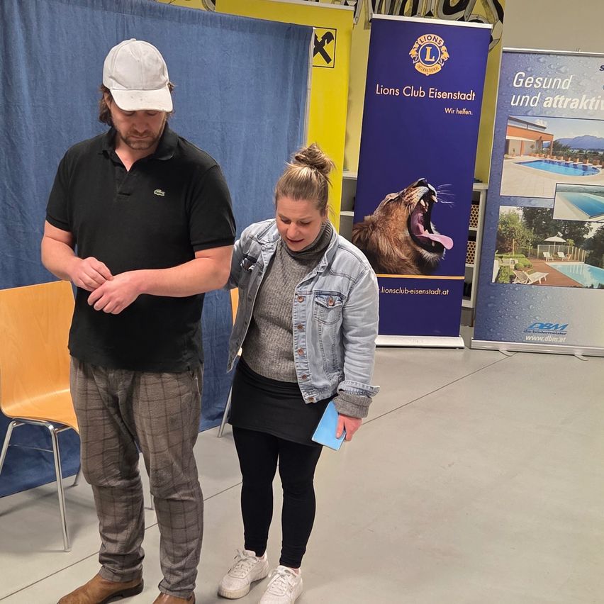A man and a woman stand in an indoor area, perhaps at an event. The man is wearing a black polo shirt and plaid pants, while the woman is wearing a denim jacket. They are both looking at something on the right. Behind them are banners with text and a lion.