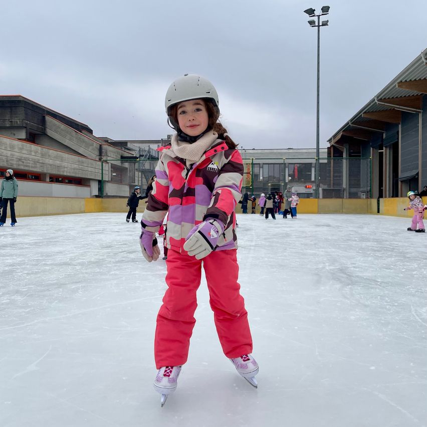 A young girl skates on an ice rink, wearing a helmet and colorful ski suit. In the background, there are other people skating and buildings with a cloudy sky.
