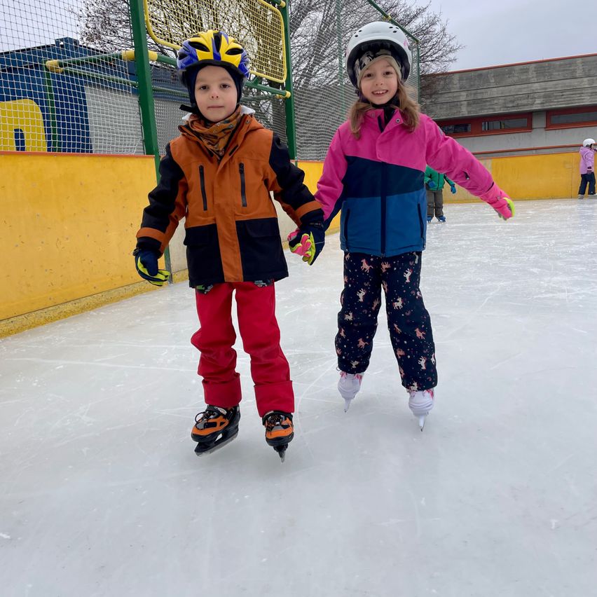 Two children, one boy and one girl, are ice skating on an outdoor ice rink. They are both wearing winter clothes, helmets, and gloves. They are holding hands. Behind them is a building with a fence.
