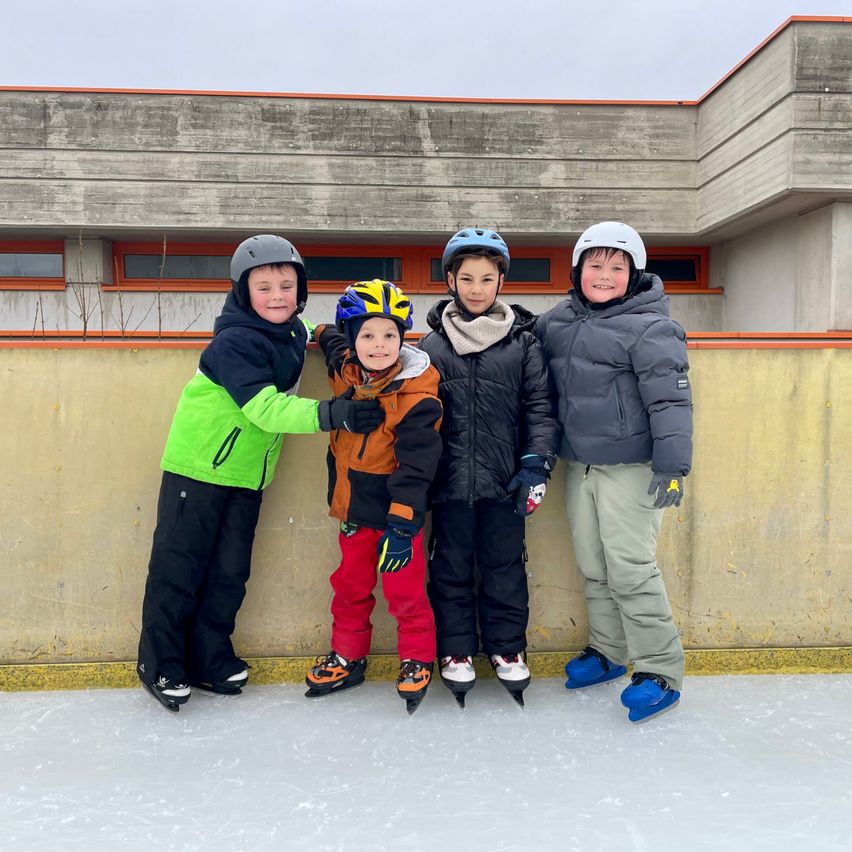 Four boys in winter gear, smiling, stand on an ice rink in front of a building with yellow and white walls. They wear helmets and gloves.