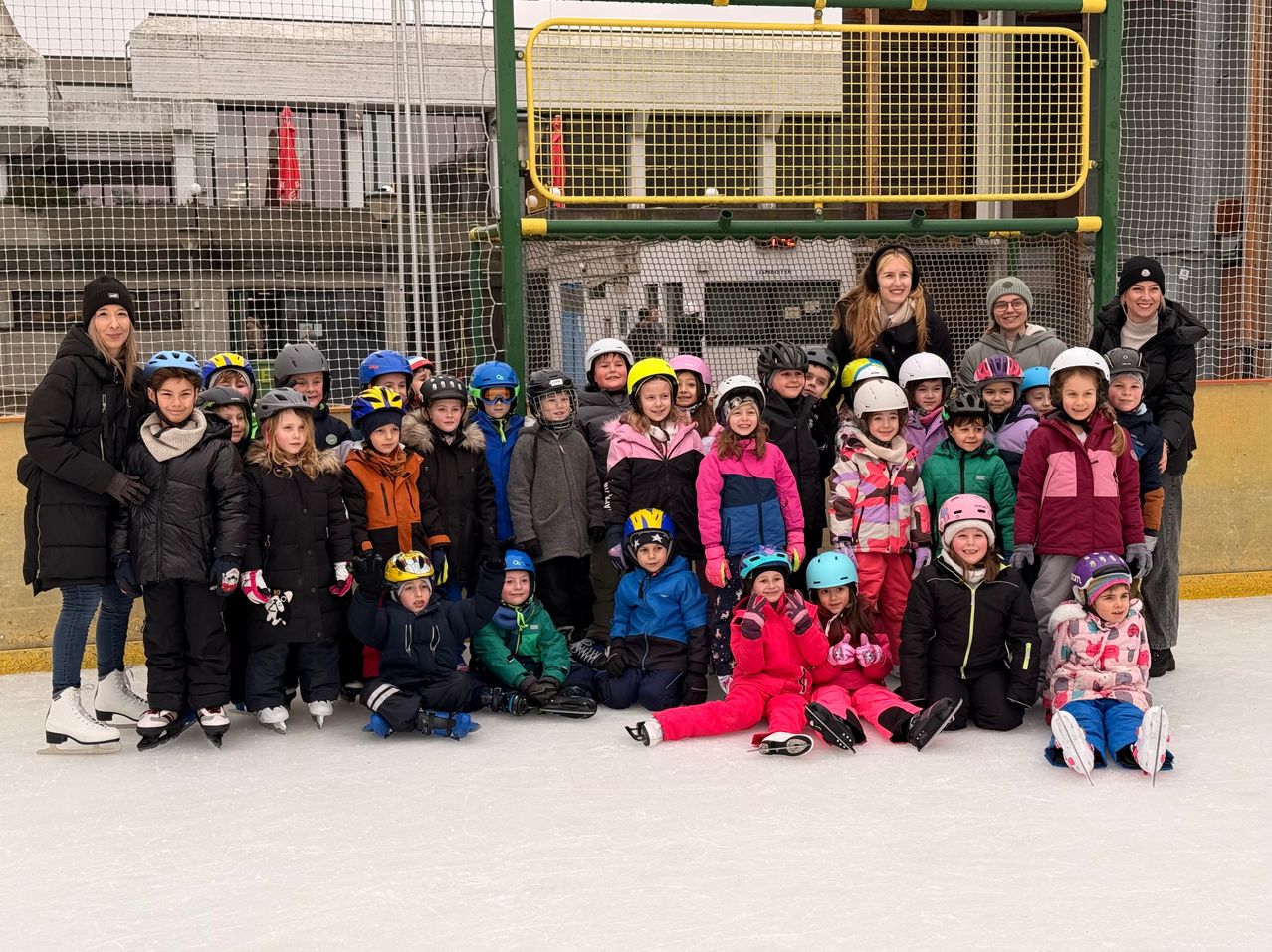 A group of children, all wearing winter clothes and helmets, pose for a photo on an ice rink. Behind them, a woman stands with a fence in the background.
