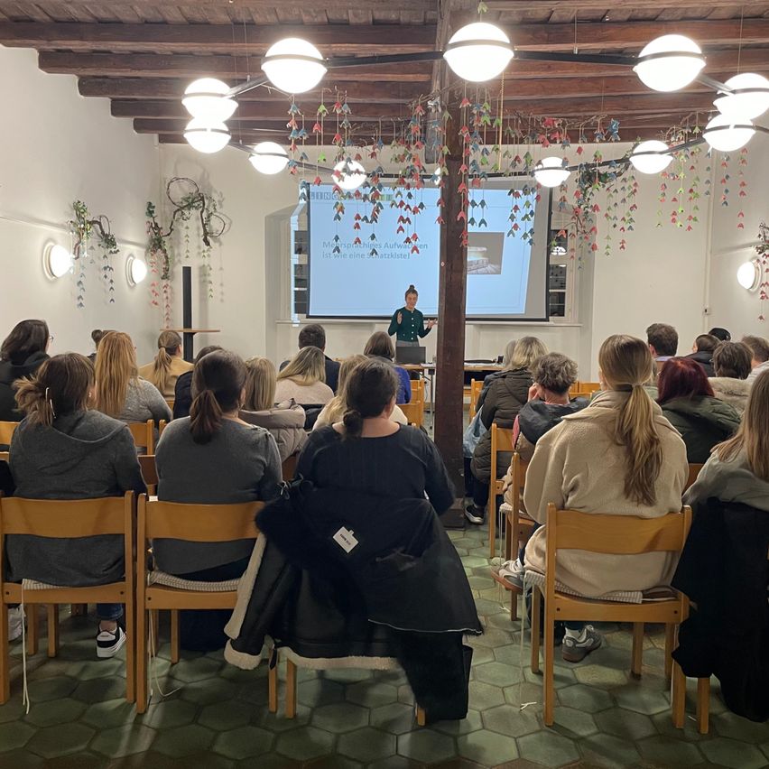 Eine Person hält eine Präsentation in einem Raum mit einer Projektionsleinwand. Eine Menschenmenge sitzt auf Stühlen. Der Raum hat Holzbalken und dekorative Lichter.